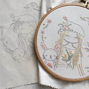 May include: Embroidered hoop art featuring a girl with long hair wearing a floral crown and holding a bunny. The girl is wearing a pink and white dress and the bunny is white with pink ears. The background is a light beige fabric.