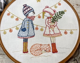 May include: Hand-stitched embroidery art in a wooden hoop. The design features two figures in winter coats and hats, with a sleeping fox at their feet. A string of lights and a star are also embroidered.