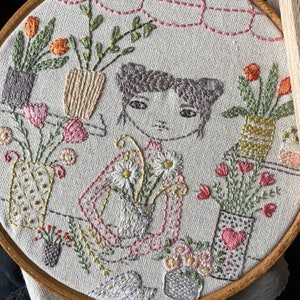 以下が含まれることがあります： 女の子、花、鉢植えの植物が描かれた、ファンシーなシーンの刺繍フープアート。デザインは、ナチュラルリネン生地にピンク、グリーン、ブラウンの色合いで刺繍されています。