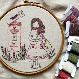 Può includere: Ricamo su telaio in legno con un design stravagante. Una figura con cappotto e sciarpa è vicino a una cassetta postale con un uccello che tiene una lettera. Il testo "PIGEON POST" è visibile. Filo da ricamo di vari colori sullo sfondo.