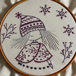 May include: Hand-stitched embroidery art in a wooden hoop. The design features a girl wearing a purple hat and cape, with white hair and snowflakes. The embroidery is on a cream-colored fabric, creating a winter theme.