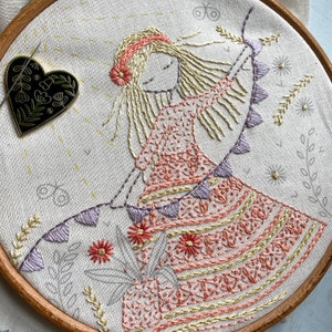 以下が含まれることがあります： 刺繍されたフープアート。長いブロンドの髪をした女の子が、ピンクとオレンジのドレスを着て、紫色の旗の紐を持っています。背景はクリーム色の布で、刺繍された花と葉が描かれています。