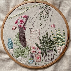 Pode incluir: Arte de bordado em aro que apresenta uma mulher com roupa estampada floral rosa e branco segurando uma planta em vaso. O fundo inclui várias plantas e borboletas em tons de verde, rosa e branco.