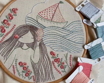 Peut inclure: Un cercle à broder avec une broderie représentant une jeune fille aux longs cheveux, une coquille et un voilier sur la mer. Le motif utilise des fils roses, bleus et bruns. Plusieurs bobines de fil à broder DMC sont visibles.