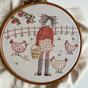 May include: Embroidered hoop art featuring a girl in a red and white dress holding a bucket, with three chickens in the background. The girl has brown hair and is standing in front of a fence.