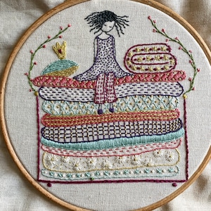 Può includere: Un telaio da ricamo circolare contenente un disegno cucito a mano di una persona con capelli scuri seduta sopra un'alta pila di cuscini colorati e decorati. I cuscini presentano varie texture, inclusi motivi floreali, geometrici e a righe.