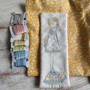 Pode incluir: Um marcador de página de tecido branco com um desenho bordado à mão de uma menina com um vestido branco com detalhes azuis em pé sobre uma pilha de livros. A menina tem cabelo loiro e olhos azuis. O tecido é de cor creme claro com uma textura sutil. O marcador de página é rodeado por um tecido amarelo com um padrão floral.