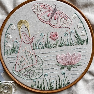 以下が含まれることがあります： 木製のフレームに刺繍されたアートワーク。ピンクのドレスを着た女の子が蝶に繋がれた紐を持っているデザインです。ピンクと緑の花柄とピンクの蓮の花も含まれています。