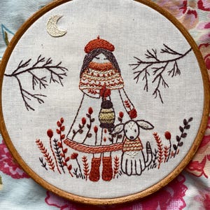 May include: Embroidered artwork in a wooden hoop, depicting a girl with a lantern and a dog in an autumnal scene. The design uses brown, orange, and cream thread on a white background. A crescent moon is stitched in the sky.