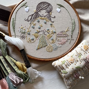 May include: A beige embroidery hoop featuring a hand-stitched design of a girl with brown hair and a floral dress. The girl is holding a bundle of yarn and there are flowers and leaves surrounding her. The hoop is on a white surface.
