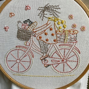 Pode incluir: Imagem bordada de uma pessoa a andar de bicicleta com uma cesta cheia de itens. A pessoa está a usar um vestido estampado em castanho e laranja e tem cabelo castanho encaracolado. A bicicleta é rosa com rodas castanhas e uma cesta castanha. O fundo é um tecido branco.