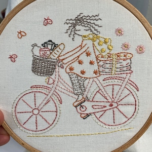 May include: Embroidered image of a woman riding a bicycle with a basket full of items. The woman is wearing a brown and yellow dress and has brown hair. The bicycle is pink and has a brown basket. The background is white.