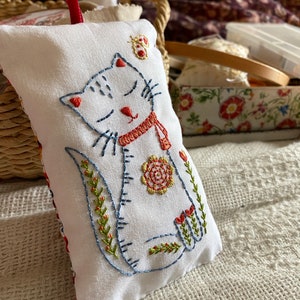 May include: White fabric pincushion with a hand-stitched design of a cat with closed eyes, a red scarf, and a flower. The cat is surrounded by green leaves and a small red butterfly.