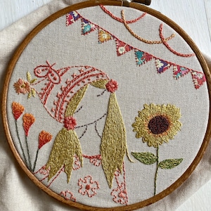 May include: A circular wooden embroidery hoop containing a fabric panel with hand-stitched thread art. The design features a person with long yellow hair wearing a patterned hat, surrounded by orange flowers, a large yellow sunflower, and colorful bunting.
