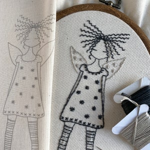 May include: An embroidery hoop featuring a hand-stitched fairy design. The fairy wears a dress with star details and striped leggings, with curly hair and angel wings. A pencil outline of the design is on a separate piece of fabric. Thread spools are visible.