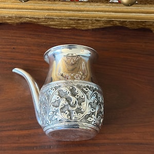 May include: A small, polished silver-toned metal vessel with a curved spout and a wide, flared rim. The lower body features an intricate, embossed floral and leaf pattern with a dark, textured background, contrasting with the smooth, reflective upper section.