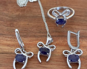 May include: A silver jewelry set featuring a ring, necklace, and earrings. Each piece has a bow-shaped design and a dark blue oval gemstone. The set is displayed on a wooden surface.