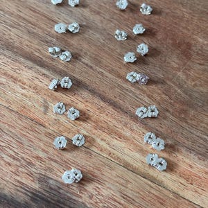 May include: A collection of silver-colored earring backs scattered on a wooden surface. The earring backs are small, with a butterfly-style design. The background is a warm-toned, wooden surface.
