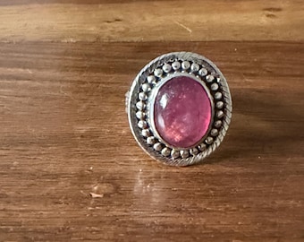 Könnte beinhalten: Ein silberner Ring mit einem großen, ovalen, rosa Edelstein in der Mitte. Der Edelstein ist von einem perlenbesetzten Rand und einem gedrehten Seilmotiv umgeben. Der Ring liegt auf einer Holzoberfläche.