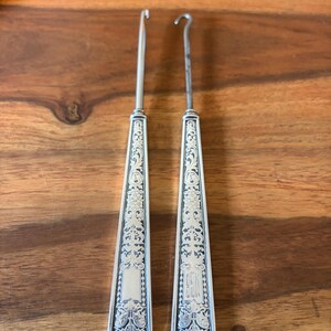 May include: A pair of antique silver crochet hooks with decorative floral and shield patterns. The handles are intricately engraved, and the hooks have a curved tip. The hooks are presented on a wooden surface.