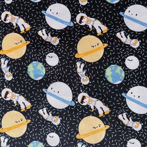 May include: Black fabric featuring a repeating pattern of planets, astronauts, and stars. The planets are white with blue rings and yellow rings. Astronauts in white suits float among the planets. The background is black with white dashes.