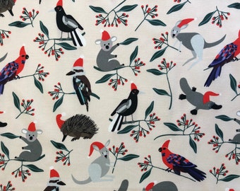 May include: A cream-colored fabric with a repeating pattern of Australian animals wearing Santa hats. The animals include a koala, a kangaroo, a platypus, an echidna, a kookaburra, a magpie, and a red-winged fairywren. The fabric is decorated with green leaves and red berries.