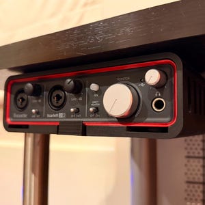 May include: Black audio interface with red trim, mounted under a dark surface. Features multiple input jacks, control knobs, and a headphone jack. The text "Scarlett 2i2" is visible.