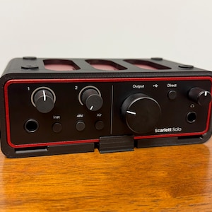 May include: Black Scarlett Solo audio interface with red accents. Features knobs, buttons, and input/output ports. The device has the text "Scarlett Solo" printed on the front. The device is designed for audio recording and production.