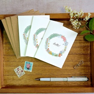 Puede incluir: Tres tarjetas de felicitación blancas con una ilustración de corona floral que incluye herramientas de jardinería y la palabra Hello, acompañadas de sobres marrones, dos sellos, un clip de metal y un bolígrafo claro con estampado floral.