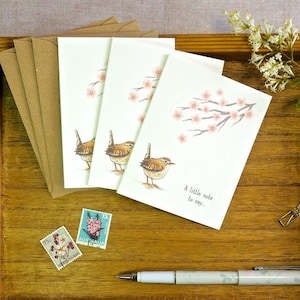 Könnte beinhalten: Drei weiße Klappkarten mit Illustrationen von braunen Zaunkönigen und rosa Kirschblütenzweigen. Die vordere Karte trägt den Text A little note to say. Enthalten sind braune Umschläge, zwei Briefmarken und ein heller Füllfederhalter.