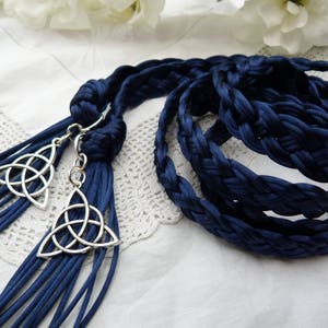 May include: A long, dark blue braided cord made of satin-like fabric, featuring two silver-toned metal triquetra charms attached to the ends. The cord is coiled and includes long, loose strands forming a tassel-like finish at the knotted base.