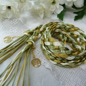 May include: A braided cord made of green, white, and gold satin ribbon with gold tree of life charms attached. The cord is braided in a complex pattern and is suitable for use as a belt or a decorative accent.
