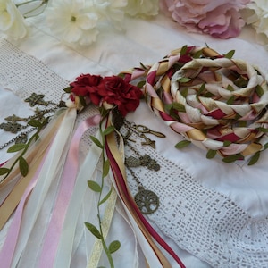 May include: A braided rope made of pink, white, and gold ribbon with green leaves woven in. The rope is tied with a red and gold ribbon and has a cluster of red flowers attached. The rope is decorated with small metal charms.