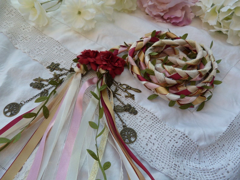 May include: A braided rope made of pink, white, and gold ribbon with green leaves woven in. The rope is tied with a red and gold ribbon and has a cluster of red flowers attached. The rope is decorated with small metal charms.
