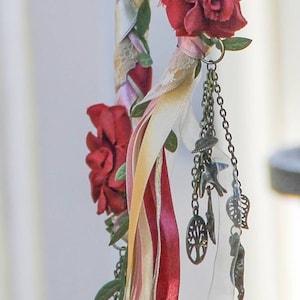 May include: A decorative wand with a braided ribbon design featuring pink, white, and red ribbons. The wand is adorned with red fabric flowers and a chain with charms, including a tree of life pendant.