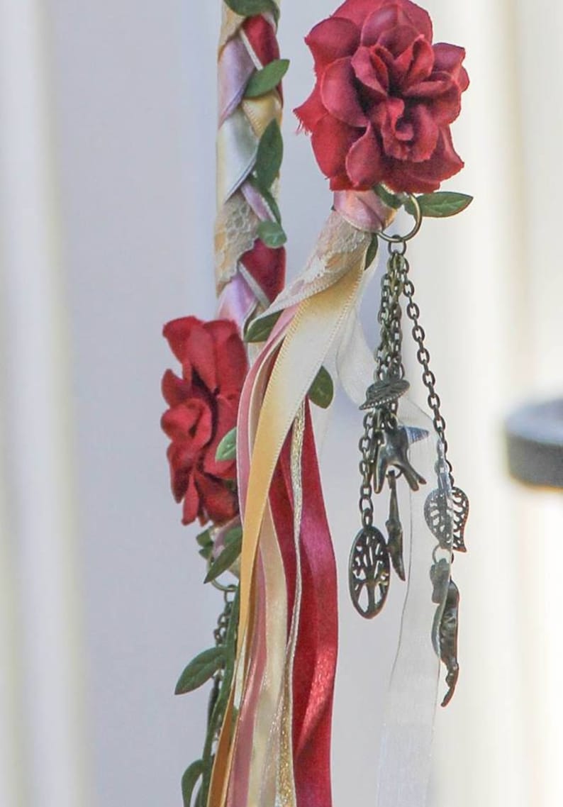May include: A decorative wand with a braided ribbon design featuring pink, white, and red ribbons. The wand is adorned with red fabric flowers and a chain with charms, including a tree of life pendant.