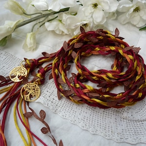 May include: A braided cord with burgundy, gold, and orange strands, decorated with brown fabric leaves and gold tree of life charms.
