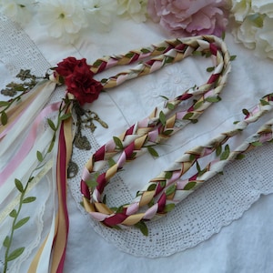 May include: A braided garland made of white, pink, gold, and burgundy ribbons with green leaves woven throughout. The garland is decorated with a cluster of burgundy fabric roses and antique-looking charms.