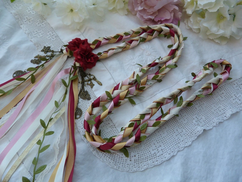 May include: A braided garland made of white, pink, gold, and burgundy ribbons with green leaves woven throughout. The garland is decorated with a cluster of burgundy fabric roses and antique-looking charms.