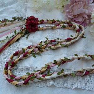 May include: A braided wreath made of burgundy, pink, cream, and gold ribbon with green leaves woven throughout. The wreath is tied with a burgundy ribbon and has a small red flower attached.