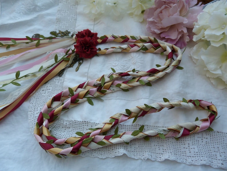 May include: A braided wreath made of burgundy, pink, cream, and gold ribbon with green leaves woven throughout. The wreath is tied with a burgundy ribbon and has a small red flower attached.