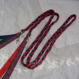 Peut inclure: Deux cordons tress&eacute;s en ruban rouge, bleu et vert avec des breloques en n&oelig;ud celtique attach&eacute;s. Les cordons sont dispos&eacute;s sur un fond de tissu blanc.