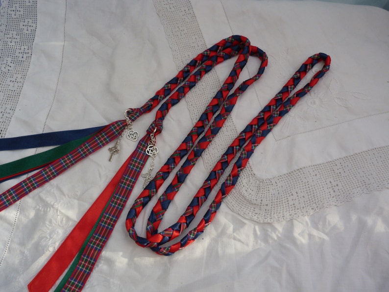 Peut inclure: Deux cordons tress&eacute;s en ruban rouge, bleu et vert avec des breloques en n&oelig;ud celtique attach&eacute;s. Les cordons sont dispos&eacute;s sur un fond de tissu blanc.