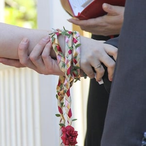 May include: A close-up of a wedding ceremony with a couple holding hands. The bride is wearing a white dress and the groom is wearing a black suit. A braided cord with red, pink, and yellow ribbons and green leaves is tied around the bride's wrist.