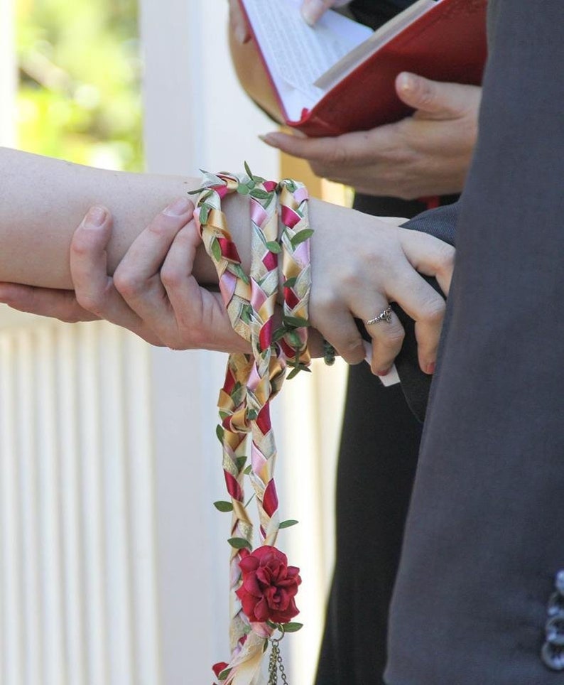May include: A close-up of a wedding ceremony with a couple holding hands. The bride is wearing a white dress and the groom is wearing a black suit. A braided cord with red, pink, and yellow ribbons and green leaves is tied around the bride's wrist.