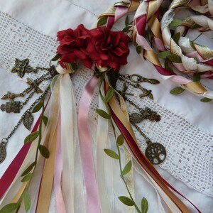 May include: A white, gold, pink, and burgundy braided rope with green leaves woven throughout. The rope is decorated with a cluster of red fabric roses and a chain of charms, including a tree, birds, and leaves.