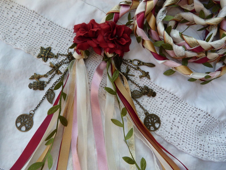 May include: A white, gold, pink, and burgundy braided rope with green leaves woven throughout. The rope is decorated with a cluster of red fabric roses and a chain of charms, including a tree, birds, and leaves.