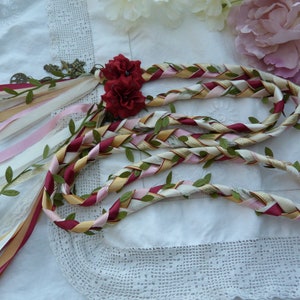May include: A braided ribbon garland with red, pink, gold, and white ribbons, interwoven with green leaves. The garland is decorated with a small, antique-looking charm and two red fabric flowers.