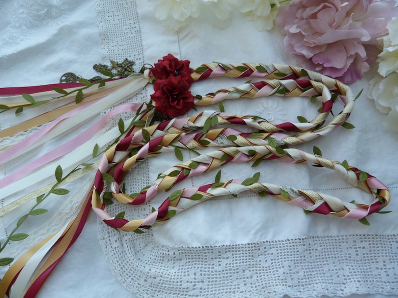 May include: A braided ribbon garland with red, pink, gold, and white ribbons, interwoven with green leaves. The garland is decorated with a small, antique-looking charm and two red fabric flowers.