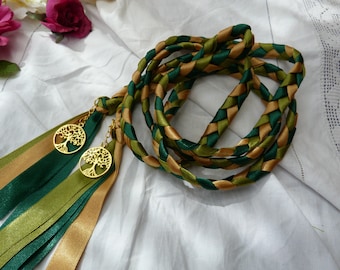 May include: A braided cord made of dark green, olive green, and gold satin ribbons. The cord features two gold-toned metal charms shaped like trees with intricate branches and roots, attached to the ends with loose, flowing ribbon tails.